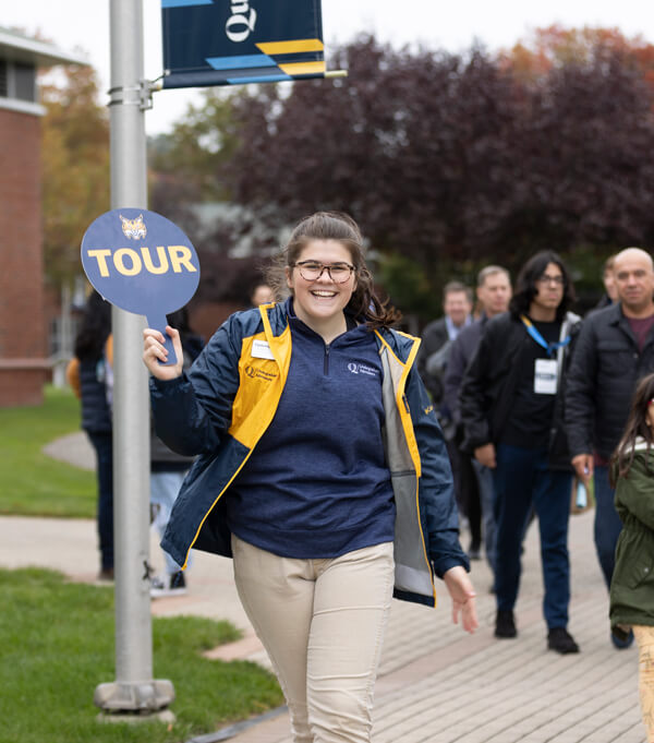 A student employee of admissions holds a tour sign and smiles as she leads a tour group